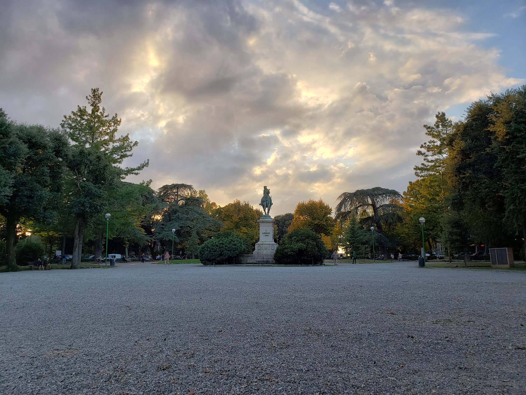 View of a gravel square surrounded by trees with a statue in the middle and clouds in the sky.